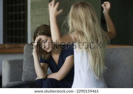Family conflict - child throwing tantrum at tired mother. Unhappy tired mother holding head, sitting on couch at home having problem with noisy naughty little daughter demanding attention.