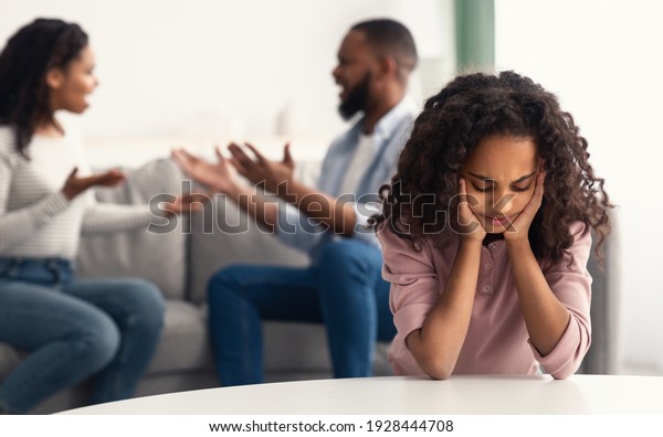 Family Conflicts. Sad little black girl covering\
ears with hands while her parents arguing in the background, upset\
child doesn\'t want to hear quarrel, stressed kid sitting alone,\
selective focus
