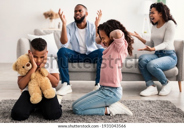 Family Conflicts. Sad little black children\
covering ears with hands while their parents arguing in the\
background, upset boy and girl don\'t want to hear quarrel, stressed\
kid sitting on the floor