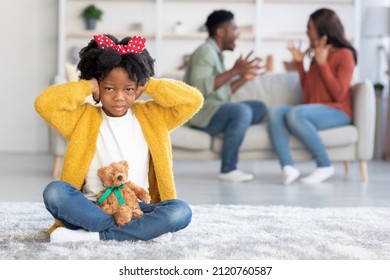 Family Conflicts. Upset Black Girl Closing Ears With Hands While Parents Arguing On Background, Sad African American Female Child Sitting On Floor Alone, Trying Not To Hear Quarrels, Selective Focus