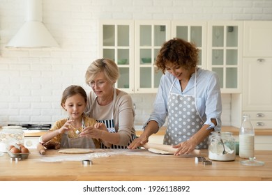 Family cooking. 3 generations women prepare bakery at home kitchen. Young female mom roll dough while elderly aged grandma teach little girl grandkid daughter press cookies using diverse metal cutters - Powered by Shutterstock