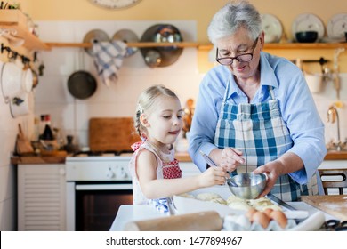 Family Is Cooking In Cozy Kitchen At Home. Grandmother And Funny Kid Are Making Italian Meal. Senior Woman And Child Girl Are Baking Bread. Cute Is Helping To Prepare Dinner. Children Chef Concept