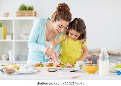 Family, Cooking And People Concept - Mother And Little Daughter Making And Decorating Cupcakes With Cream Frosting At Home Kitchen