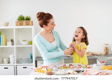 Family, Cooking And People Concept - Mother And Little Daughter Making And Decorating Cupcakes With Sprinkles At Home Kitchen