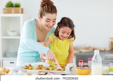 Family, Cooking And People Concept - Mother And Little Daughter Making And Decorating Cupcakes With Cream Frosting At Home Kitchen