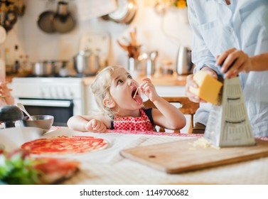 Family cooking pizza in kitchen. Mother and daughter preparing homemade italian food. Funny little girl is helping woman, eating and tasting cheese and ingredients. Children chef concept.