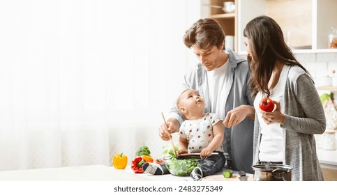 A family is cooking together in the kitchen. The father is holding a baby who is stirring a bowl of salad with a wooden spoon. The mother is standing next to them, holding a tomato, copy space - Powered by Shutterstock