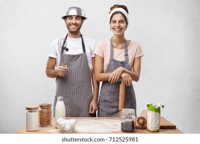 Family Couple Cooking At Home, Baking Cake, Having Happy Expressions. Pretty Female With Dirty Face Holding Rolling Pin In Hands Standing Near Her Attractive Husband Who Has Fun Posing With Whisk