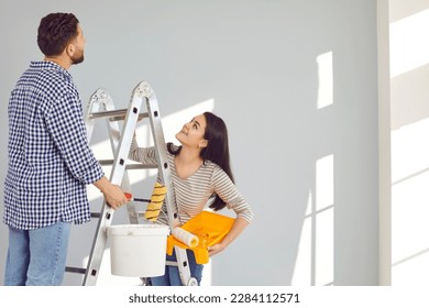Family couple doing renovations, painting walls and whitewashing ceiling at home. Happy young man and woman standing on step ladder, holding paint can, tray, and rollers, looking up, and smiling