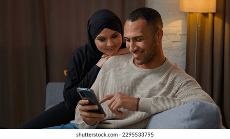 Family couple multiethnic interracial family African American man chatting on mobile phone muslim woman wife cuddle to husband use cellphone shopping online together watch funny internet video at home - Shutterstock ID 2295533795