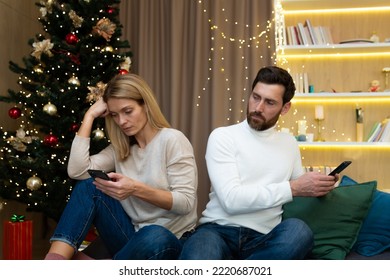 Family couple quarreled on Christmas sitting at home on sofa, husband and wife not talking to each other, holding phones in hands browsing social networks, near Christmas tree.