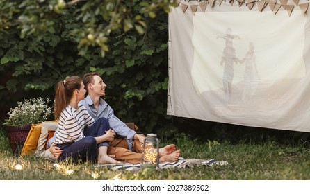 Family Couple  Sitting On Blanket With Pillows And Watching Movie Projected On Cloth In Summer Evening In Yard