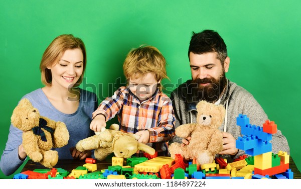 Family with curious faces hold teddy bears\
near colored construction blocks. Childhood and playing concept.\
Mom, dad and kid in playroom. Man with beard, woman and boy play on\
green background.