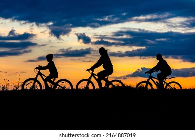 Family Cyclist And Bicycle Silhouettes On The Dark Background Of Sunsets. Mom And Her Children Ride Bicycles Against The Backdrop Of Sunset.