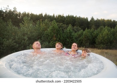 Family Dad, Mom And Two Kids Rests In A Hot Tub In Autumn Cold Weather  Against The Background Of Green Pine Forest.  Weekend Getaway At Glamping.