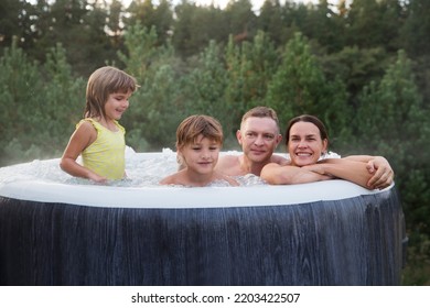 Family Dad, Mom And Two Kids Rests In A Hot Tub In Autumn Cold Weather  Against The Background Of Green Pine Forest.  Weekend Getaway At Glamping.