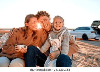 A family with a daughter sitting together and drinking coffee or tea. In the background is a car with an open trunk. Family traveling. Travel, vacation and tourism concept. - Powered by Shutterstock