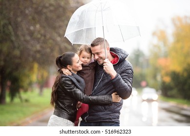 Family With Daughter Under The Umbrellas Walk On Rainy Day.