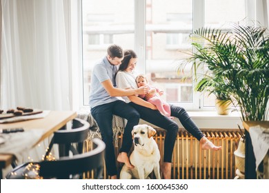 Family With Daugther And Dog Sitting Near The Big Window At The Modern Apartment.