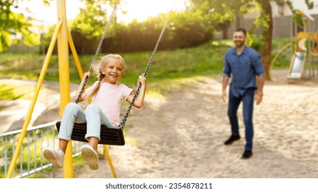 Family day. Happy girl swinging on swings having fun with father on outdoor playground in park, panorama, selective focus on smiling schoolgirl while dad standing on background - Powered by Shutterstock