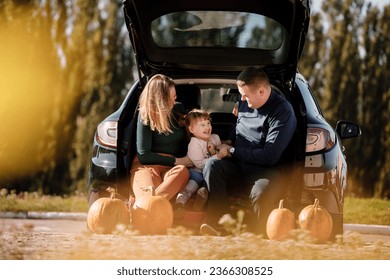 Family day. Happy mom, dad and little child daughter sit at car trunk, look at camera at autumn park with pumpkins, enjoy spend time together at weekend. Parental care and happy carefree childhood. - Powered by Shutterstock