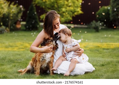 Family Day, Mother's Day. Beautiful Smiling Young Mom And Baby Daughter Cuddling Happy Domestic Dog On The Backyard Lawn.Mother With Child Girl Are Having Fun With Pet Outdoors On Summer Holiday