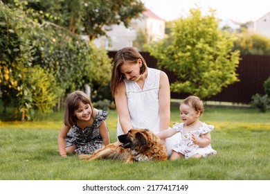 Family Day, Mother's Day. Beautiful Smiling Young Mom And Two Child Daughters Cuddling Happy Domestic Dog On The Backyard Lawn. Idyllic Family Having Fun With Pet Outdoors On Summer Holiday