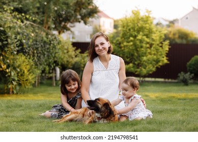 Family Day, Mother's Day. Beautiful Smiling Young Mom And Two Child Daughters Cuddling Happy Domestic Dog On The Backyard Lawn. Idyllic Family Having Fun With Pet Outdoors On Summer Holiday