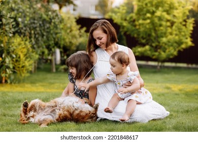 Family Day, Mother's Day. Beautiful Smiling Young Mom And Two Child Daughters Cuddling Happy Domestic Dog On The Backyard Lawn. Idyllic Family Having Fun With Pet Outdoors On Summer Holiday