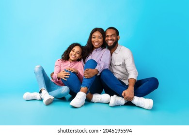 Family Day, Parenthood And Childhood Concept. Smiling Funny Young Black Parents Mom Dad Sitting On The Floor With Child Teen Girl, Hugging Daughter Isolated On Blue Color Background, Studio Portrait