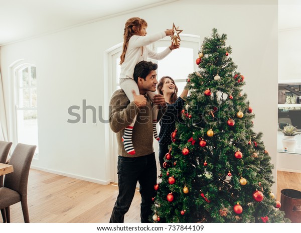 Family
decorating a Christmas tree. Young man with his daughter on his
shoulders helping her decorate the Christmas
tree.