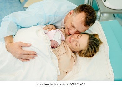 Family In The Delivery Ward. In Hospital Tired Mother Holding Newborn Baby Girl, Supportive Father Lovingly Hugging Baby And Wife.