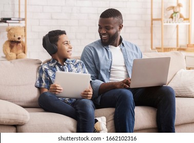 Family Devices. Joyful Black Father And His Son Using Digital Tablet And Laptop While Resting On Couch Together, Browsing Internet, Free Space - Powered by Shutterstock