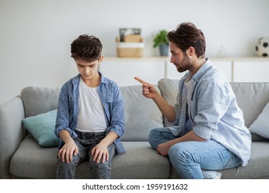 Family Discipline Concept. Grumpy Dad Scolding His Offended Son, Teaching His Kid Tolerance, Respect, Well-behaving, Sitting Together On Sofa At Home Interior