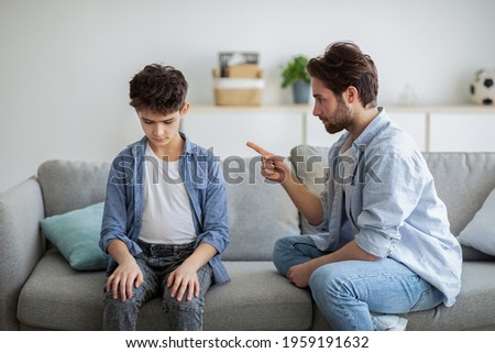Family discipline concept. Grumpy dad scolding his offended son, teaching his kid tolerance, respect, well-behaving, sitting together on sofa at home interior