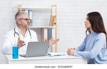 Family Doctor Consultation And Diagnostics. Serious Senior Man In White Coat And Glasses Speaks To Woman Patient At Table With Sanitizer And Laptop In Office Interior, Panorama, Empty Space, Side View