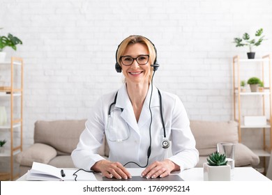 Family Doctor Consults Remotely. Smiling Woman Doctor In Headphones Sits At Table With Laptop In Living Room Interior