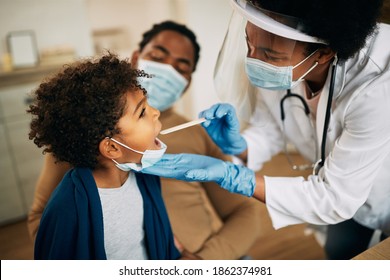 Family doctor examining throat of a small black boy while visiting him at home during coronavirus pandemic. 