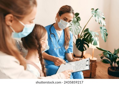 Family doctor at home in a medical protective mask examining a child with her mother sitting on the sofa in the living room. Patient examination, health care and medicine. - Powered by Shutterstock