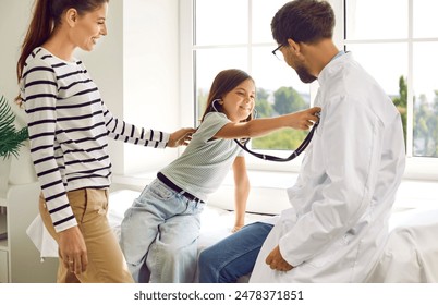 Family doctor lets kid patient have fun in his office. Mother and child visit doctor together. Happy little girl puts stethoscope to chest of her pediatrician. Health care, medical check up concept - Powered by Shutterstock