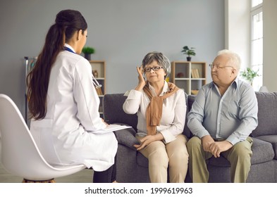 Family doctor listening to senior patients' health concerns and troubles during home visit. Old couple sitting on couch in living-room telling about migraine symptoms and asking for pain relief advice