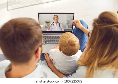 Family Doctor Online. Young Family With A Son Makes A Video Call Via Laptop To A Pediatrician For Advice And Counseling. Female Doctor Waving Hand Greeting Patients. Top View From Behind.