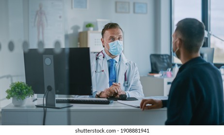 Family Doctor In Protective Mask Is Reading Medical History Of Young Male Patient And Speaking With Him During Consultation In A Health Clinic. Physician In Lab In Front Of Computer In Hospital Office