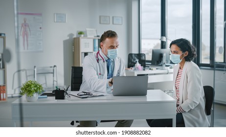 Family Doctor In Protective Mask Is Reading Medical History Of Female Patient And Speaking With Her During Consultation In A Health Clinic. Physician In Lab In Front Of Computer In Hospital Office.