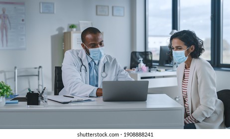 Family Doctor In Protective Mask Is Reading Medical History Of Female Patient And Speaking With Her During Consultation In A Health Clinic. Physician In Lab In Front Of Computer In Hospital Office.