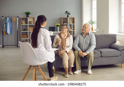 Family Doctor Visiting Patients At Home. Young Physician Listening To Married Senior Couple. Happy Mature Husband And Wife Sitting On Sofa In Living-room And Talking To Nurse Or General Practitioner