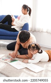 Family Does Homework At Home. Dad Helps Daughter On House's Floor. Mom Sits On Blue Couch On The Back. Kid Draws Picture On Paper.