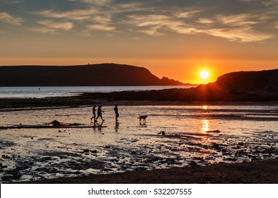 A Family With Dog In Silhouette Walking On A Beach At Daymer Bay In Cornwall England.  The Setting Sun Is Reflecting On The Wet Sand And The Sea In The Camel Estuary.