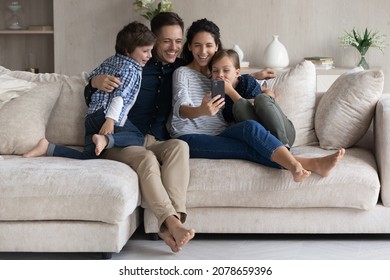 Family Enjoy Funny Mobile App Relaxing On Couch. Latin Couple And Preschool Children Use Smartphone Watch Videos On Internet Laughing Have Fun Spend Weekend At Home With Online Amusing Virtual Content