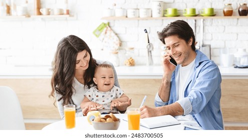 A family is enjoying breakfast together in their kitchen. The mother is sitting with her young child, who is smiling and happy. Father is sitting at the table, talking on his phone and taking notes - Powered by Shutterstock